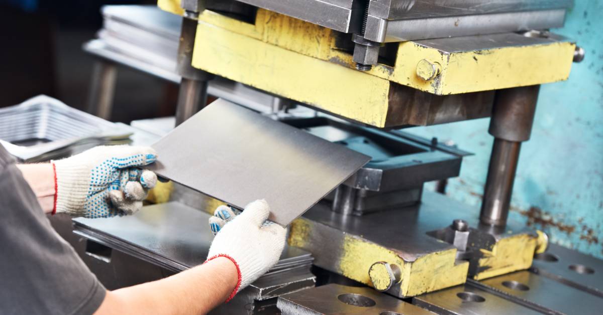 An industrial worker wearing thick gloves while loading a sheet of metal into a metal press machine in a factory.