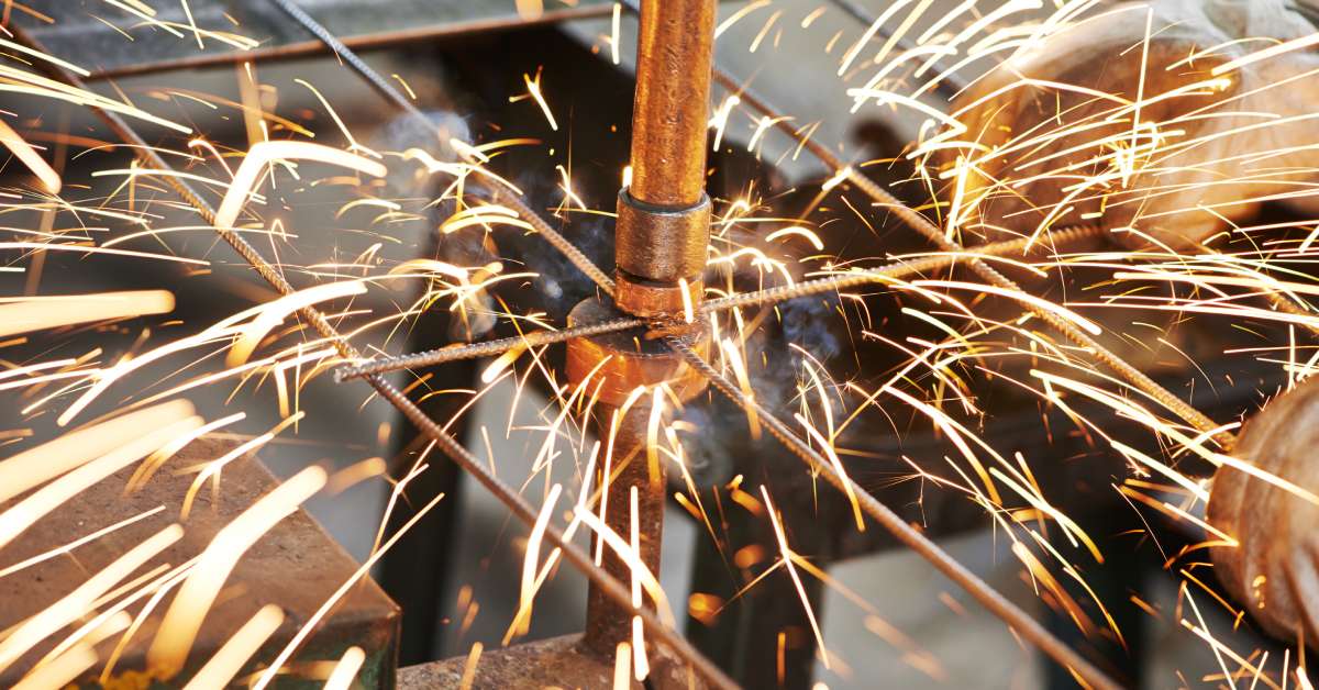 An industrial worker performing a spot welding task with a metal fence, wearing a pair of heavy work gloves.