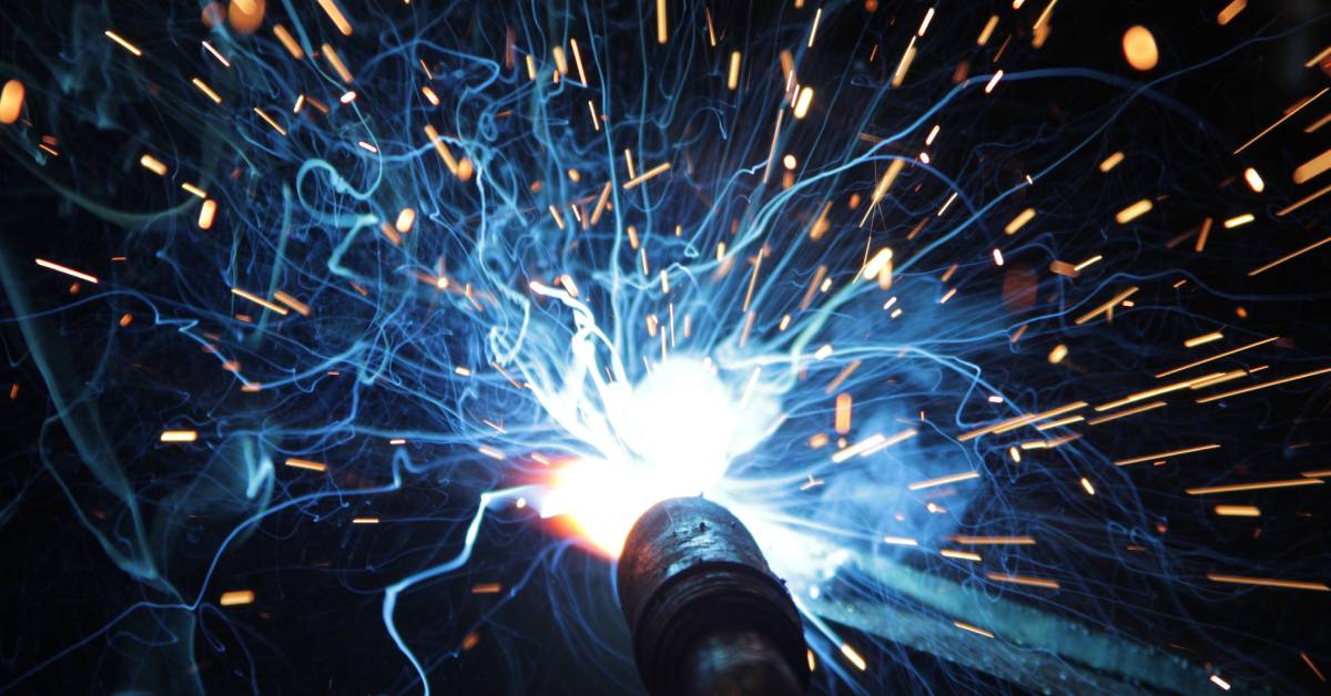 A close-up of a MIG welding torch welding a metal piece in an industrial facility. There are bright sparks flying everywhere.