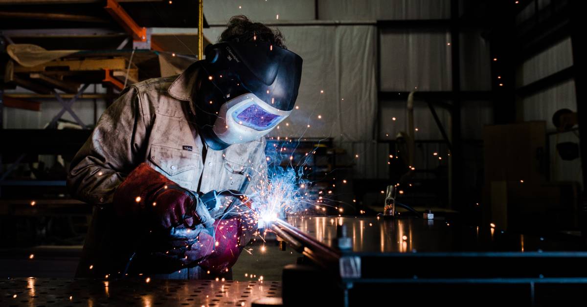 An industrial worker wearing coveralls and thick red gloves while using a MIG welding torch in a manufacturing facility.