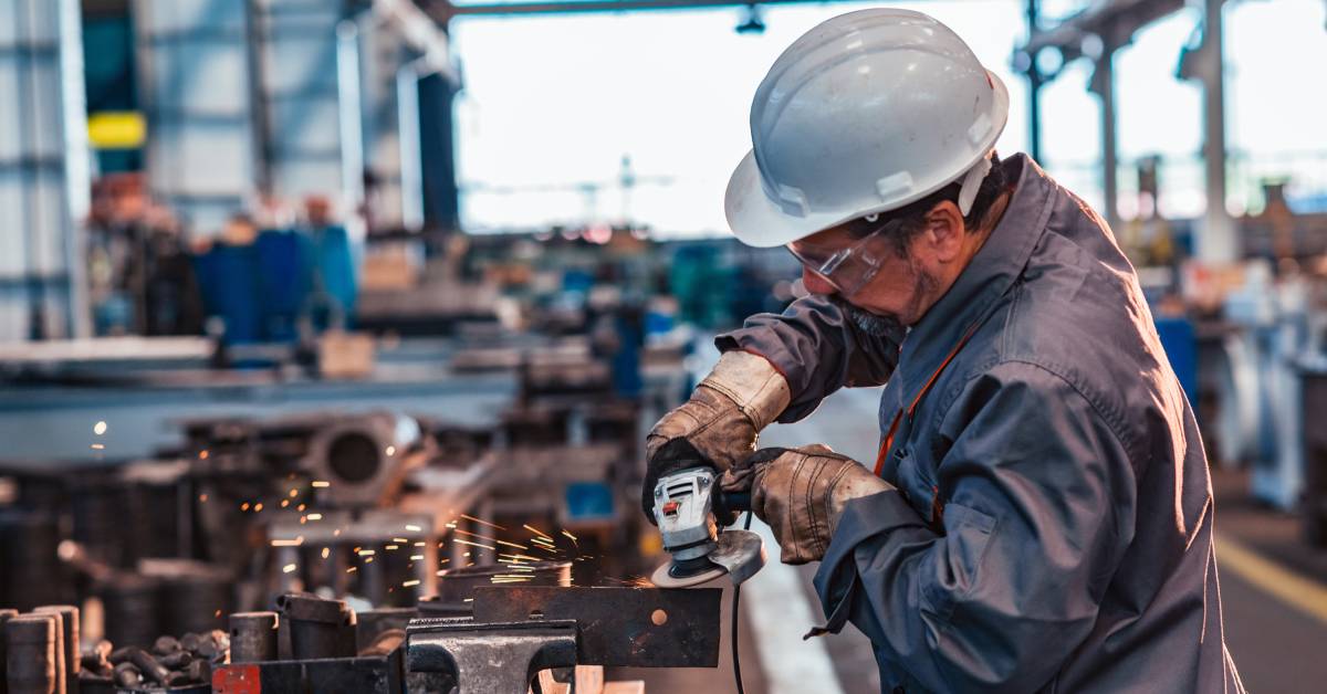 An industrial worker with a handheld grinding machine grinding down a bit of metal held in a table vise.