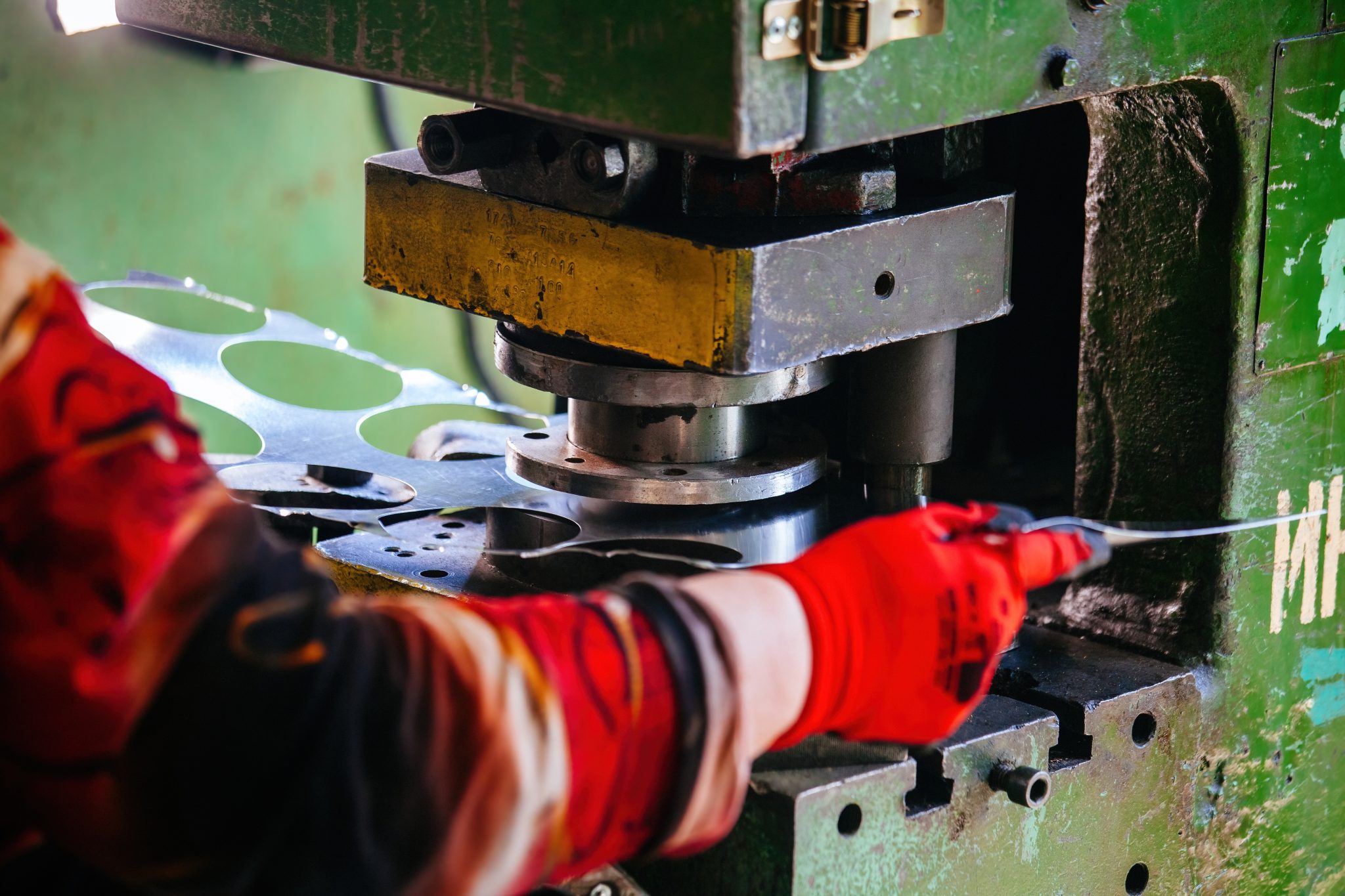 A person in red gloves skillfully operates a green stamping machine, which cuts circular shapes from a sheet of metal.