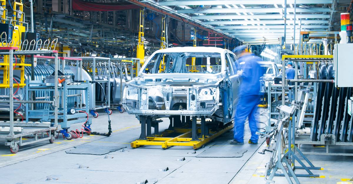 Employees working in an automotive manufacturing facility with the chassis of a sedan on a yellow production line.