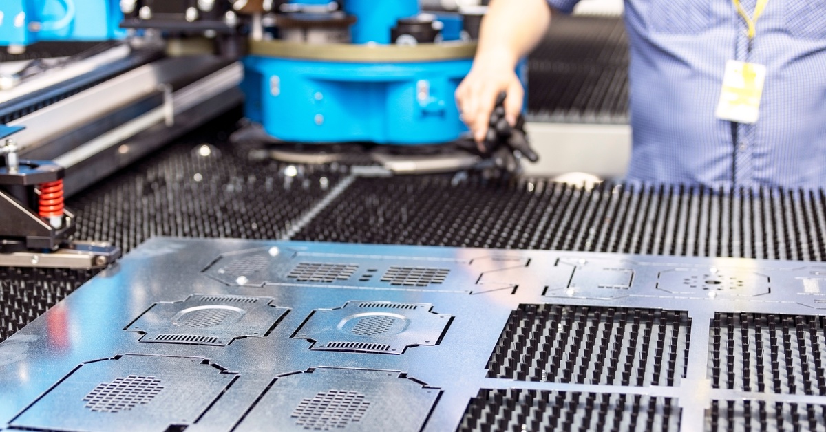 A sheet of silver metal with intricate cuts and holes in the metal lays on a black surface with a person and machines nearby.