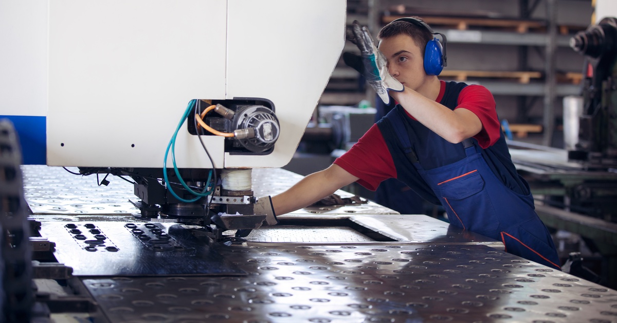 A man wearing blue noise cancelling earmuffs as he stands by a large metal sheet and has a hand on a mental press machine.