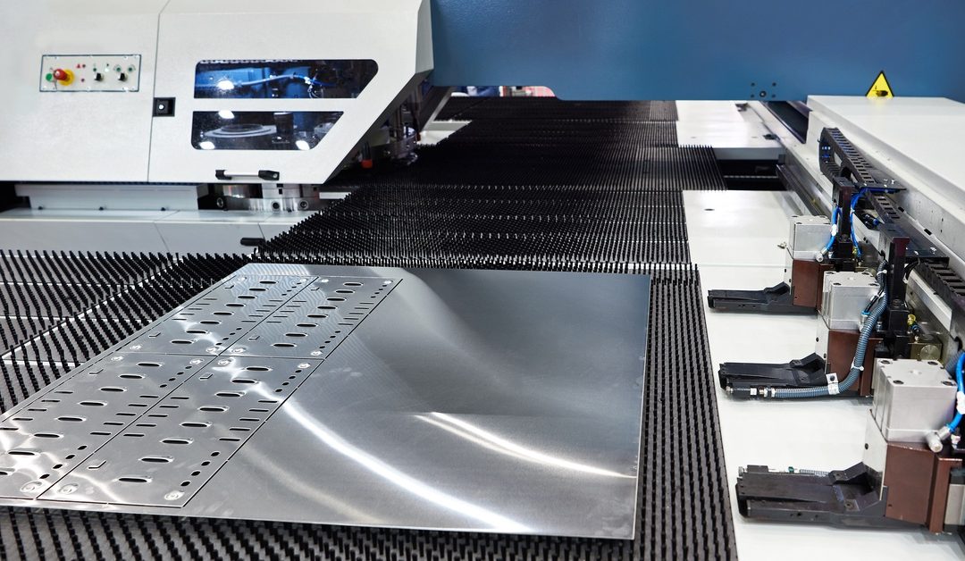A flat piece of steel lays on a black surface with machinery nearby. There are round holes and rectangular cuts in the metal.
