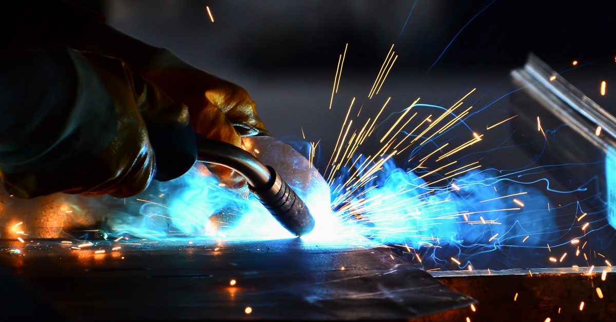 A pair of gloved hands hold a welding torch as sparks fly and blue smoke plumes around the workbench.