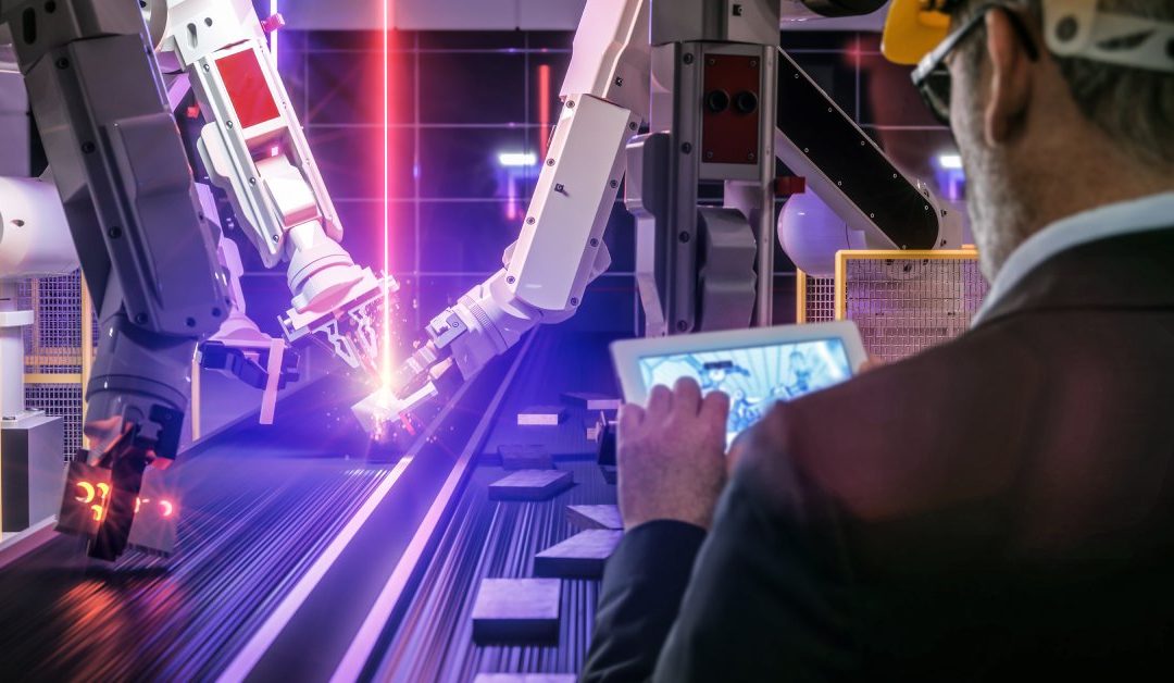 A man wearing a hard hat looks at a tablet while standing beside a conveyor system that uses robotic welding arms.
