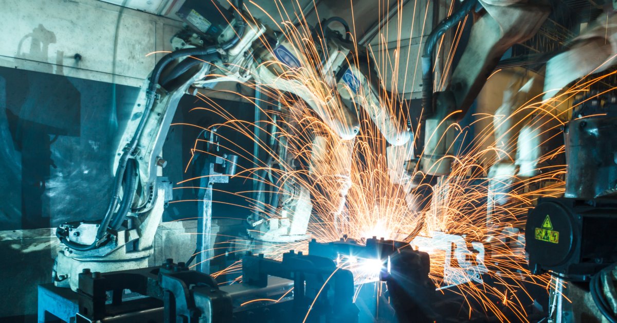 Orange sparks fly off of a piece of metal as two robotic welding arms work inside a dimly lit factory.